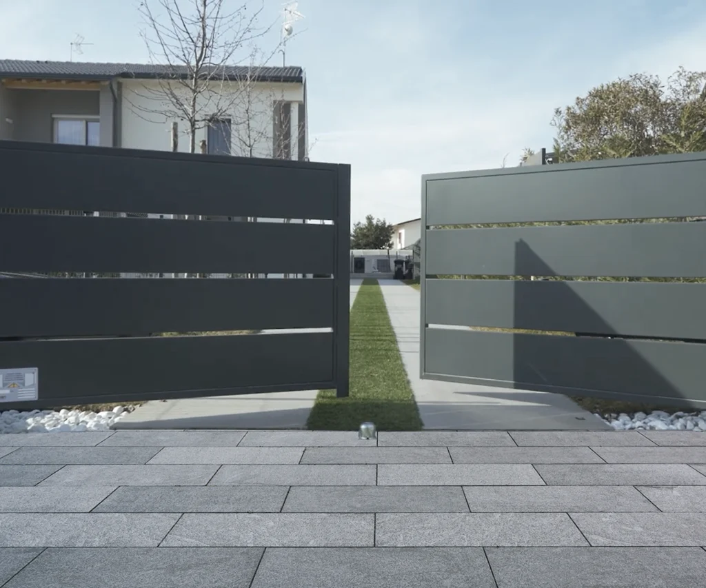 Moderne, offene Tore aus Metall führen zu einer grünen Wiese und einem Weg, umgeben von einem modernen Gebäude.