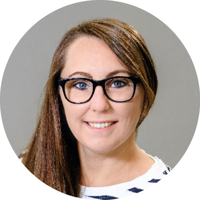Porträt einer Person mit langen, braunen Haaren und Brille, lächelnd vor einem grauen Hintergrund.